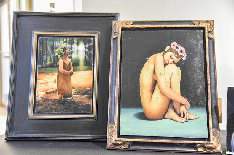 Two framed oil paintings: a girl in a forest stream and a seated woman with a flower crown
